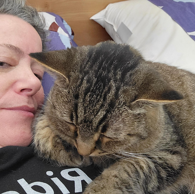 A fuzzy brown tabby naps clinging to me, a pink-faced white woman in a black t-shirt lying in bed. His closed eyes, relaxed ears, and floppy whiskers show how very comfortable and calm he is on me. One front paw is tucked under him, and the other one stretches out in front of him.