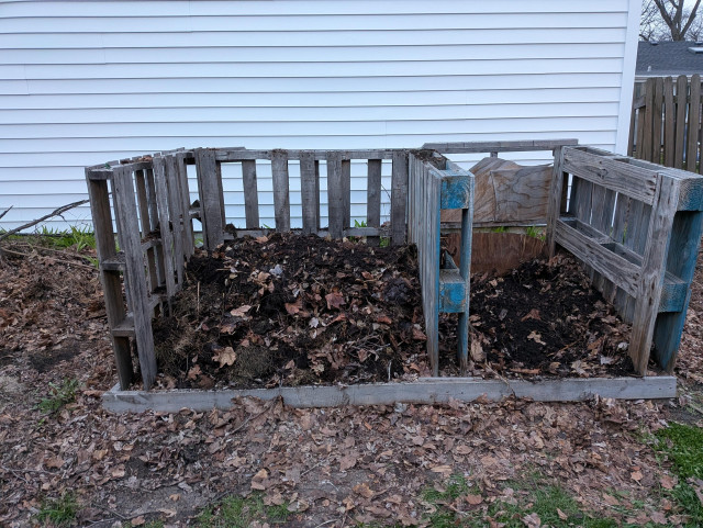 An outdoor, "cold pile" style compost bin, consisting of four pallets on end + a number of wood and plywood pieces at the foot and across the back. The construction forms an open-fronted, open-top box, with two areas inside, partitioned by one of the pallets. 

Inside the left half is a sizable mound of freshly turned compost. The right half is mostly empty, having just been moved right-to-left.
