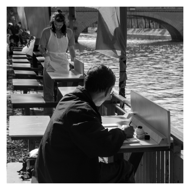 Daylight square format monochrome photography.

Foreground: a middle aged man sitting on a table drawing on a booknote, further away a young man painting a table, out of focus in background a child in a stroller.