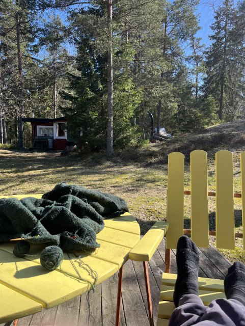 En stickning ligger på ett gult trädgårdsbord. En person sitter bredvid bordet men man ser bara fötterna (i strumplästen) som är upplagda på en stol. I bakgrunden en gräsmatta, träd med blå himmel bakom och en liten röd bod med vita knutar.