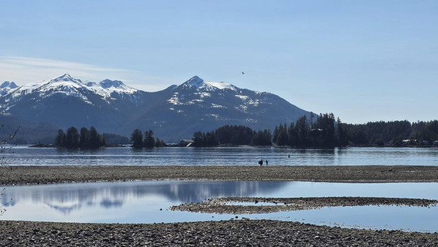 The view across the channel at low tide, the snowy mountains in the far background and reflected (Impressionistically) in a tidal pool. There are 2 figures in mid-pic, walking along a tidal promontory. 