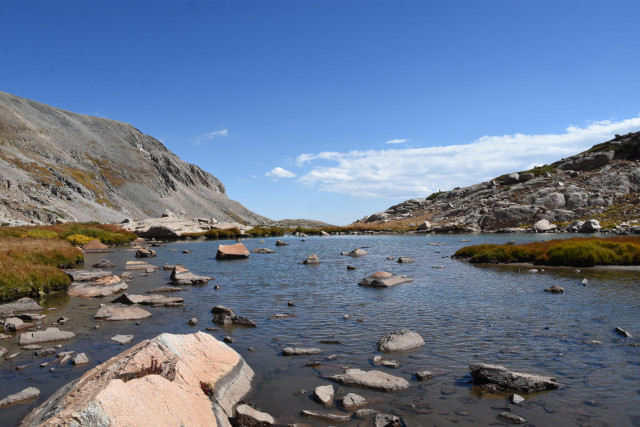 A myriad of rocks  and small islands covered in low-growing plants stick out from a shallow alpine lake. Rocky ridges jut up on either side. 