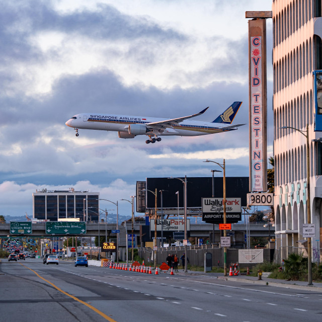 A Singapore Airlines Airbus A350 bearing registration number 9V-SJH descends over Sepulveda Blvd on approach to LAX.  The sky is cloudy with some billowing cumulus clouds, there are buildings and billboards lining the right side of the street.  There are cars on the street, and traffic cones.