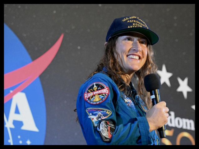Photo of NASA astronaut Christina Koch holding a microphone. She is wearing a blue jump suit with mission patches and a navy blue baseball cap, and standing in front of a large screen with NASA and other logos.
