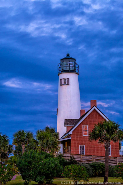 A white lighthouse with a black lantern room stands tall against a dramatic, cloudy blue twilight sky next to a two-story red brick house. Lush green tropical foliage and palm trees frame the foreground, while a wooden boardwalk leads toward the structures.