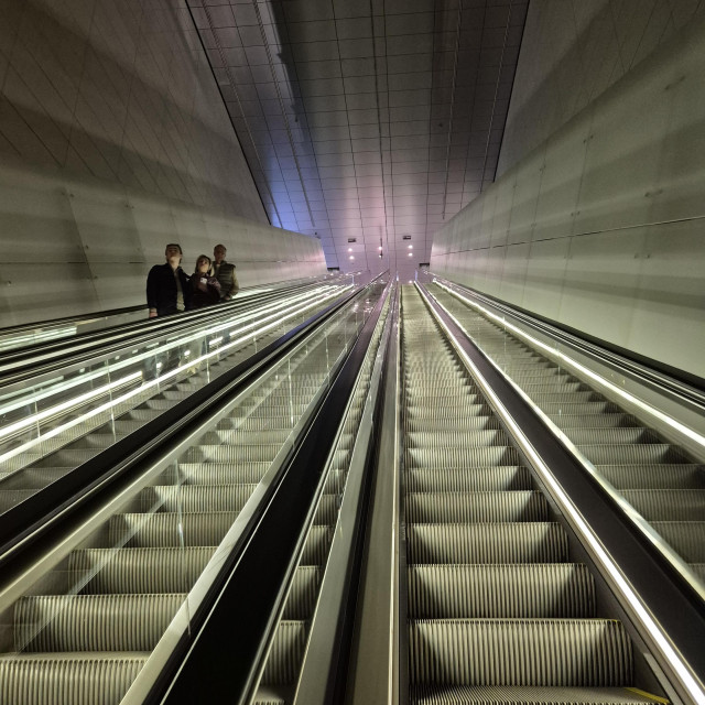 A very long empty escalator going up with a few people standing on the one going down next to it. 