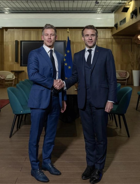 Peter Magyar in blue suit, shakes hand of Macron in front of long table with modern blue chairs, in a modern wood-paneled conference room.