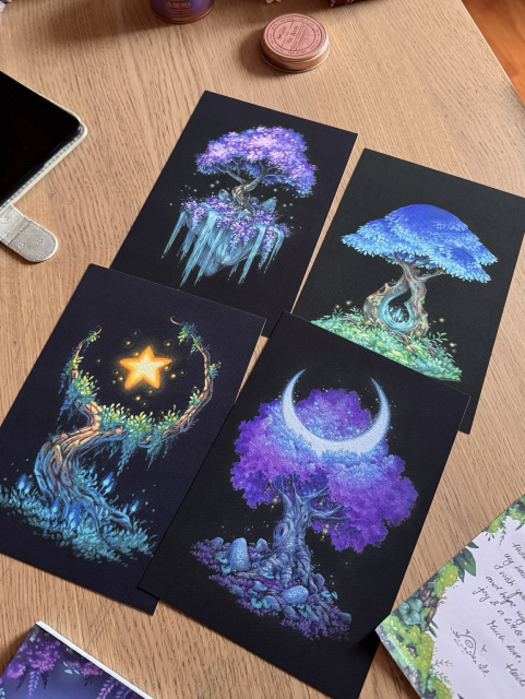 A photo of four prints of fantastic trees on black paper, placed on a wooden desk and surrounded by bits of notepads, an iPad, and the lid of a scented candle. Two of the trees feature purple foliage, one blue, and one is simply overgrown with moss and mushrooms.
