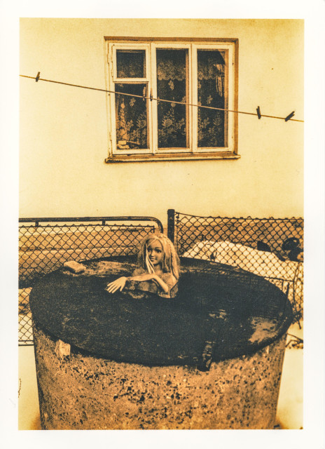 A warm-toned lith print captures a quietly surreal backyard scene. In the foreground, a concrete well ring is covered with a rusted metal lid. Resting on top of it is the upper half of a plastic doll—its pale face and disheveled hair emerging from the surface, one arm draped casually over the edge as if it has paused mid-movement.

Behind it, a simple wire fence stretches across the frame, separating the foreground from a modest house wall. A single window with patterned curtains looks outward, while a clothesline cuts diagonally through the composition, held by a few empty clothespins.