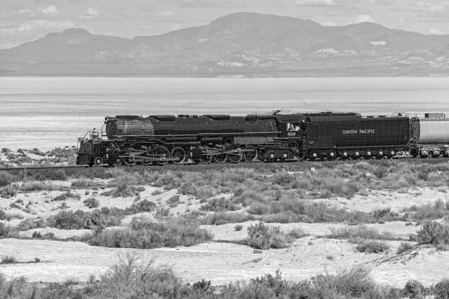 A black and white landscape photo of a very large steam engine. The train is heading toward the left side of the photo. In the foreground is some spotty clumps of sagebrush. On the opposite side of the train is a vast expanse of dry lake bed. In the far distance is a mountain range. The steam engine has two sets of four drive wheels and the boiler part is very long. The tender is attached behind the engine and printed on the side it says, "Union Pacific." A man can be seen leaning out the opening in the cab checking the track ahead. 