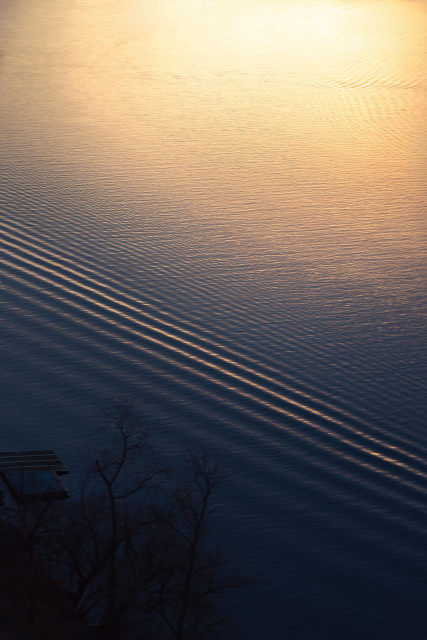 photo of a view on the Vltava river in Prague. the photo was taken at sunset. gentle waves reflect in dark blue and yellowish tones, creating stripes and patterns on the water