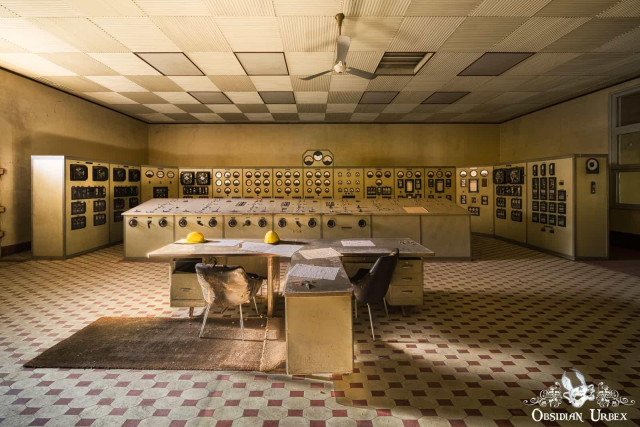 An abandoned control room featuring a large console, two desks with chairs, and a checkered floor. Two yellow hard hats rest on one desk.