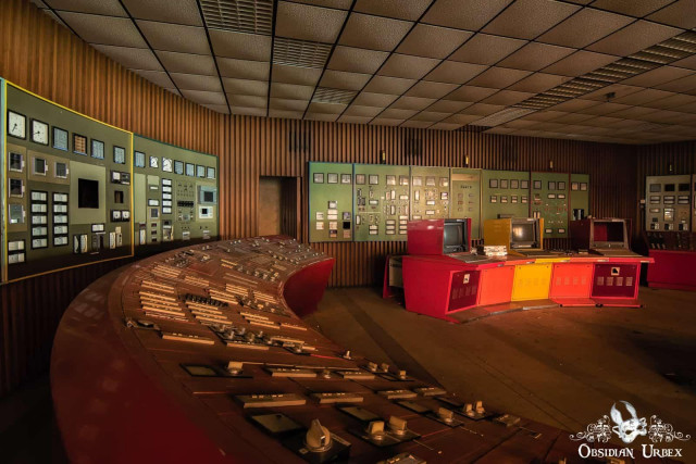 An abandoned power plant control room features a large curved red console and detailed wall-mounted control panels.