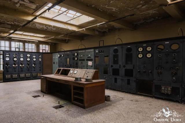 An abandoned power plant control room features a large wooden desk in the foreground and two walls of intricate, old control panels.