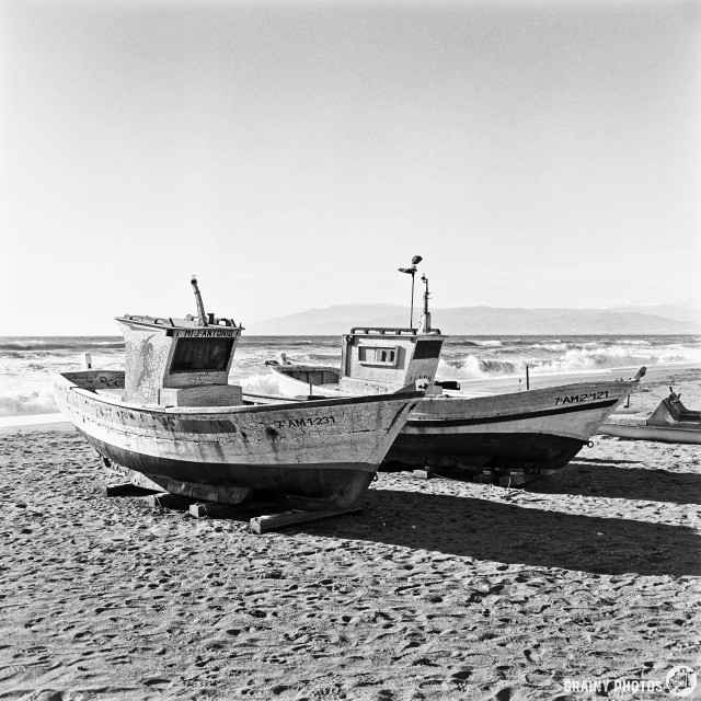 Two weathered fishing boats resting on a sandy beach, with waves crashing in the background and a clear sky. The scene captures the calm yet rugged beauty of coastal life.
