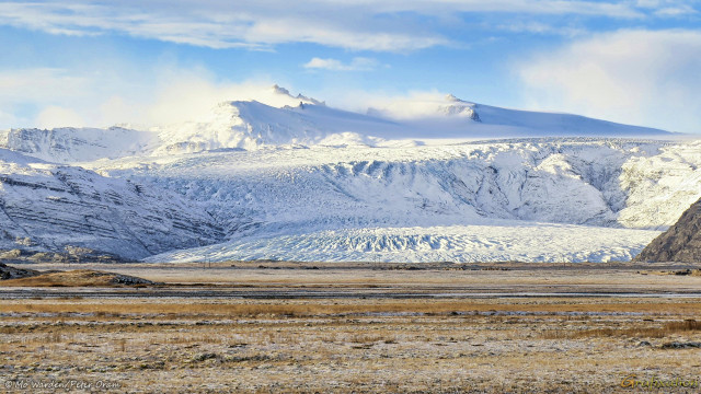 A colour photo of a winter landscape. The sky is clear but with some light cloud, and the low sunlight is from the left. The foreground is brown scrub and rocks scattered across a level surface. In the centre of the shot is an impressive glacier, sloping down in steps and covered in snow. Some heavy cracking has created huge crevasses along the lines where the angles change. At the summit are pinnacles of mountains which are mostly buried beneath the ice, snow-capped. Wind from the right is blowing clouds of snow across the surfaces, forming white clouds in the lee of the peaks and into the sunlit sides.
