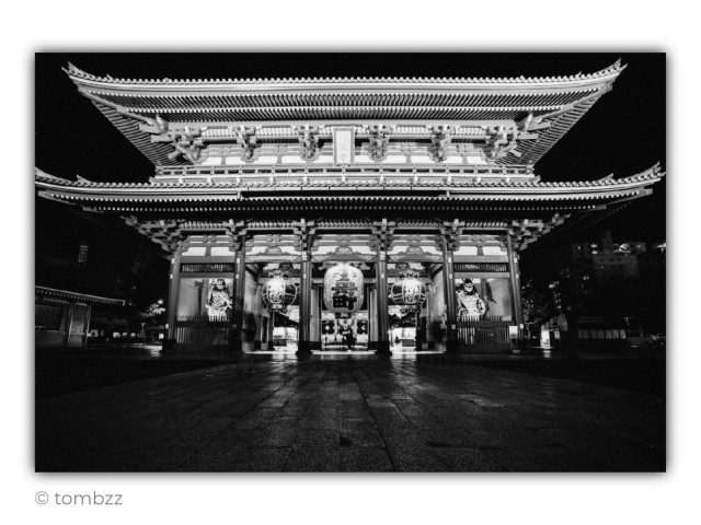 A black and white, wide-angle night photograph depicting the monumental Hōzōmon Gate, leading to the Sensō-ji Temple in the Asakusa district of Tokyo. The structure features an intricate, multi-tiered roof design with traditional ornaments and brackets, which are dramatically illuminated against the dark sky. Three large chōchin lanterns hang in the lower part of the gate – the central one is the largest, flanked by two smaller ones. Behind the grilles in the side niches, statues of the Niō guardian deities are visible. The ground-level perspective emphasizes the massive scale of the architecture, while the wet pavement of the square in front of the gate subtly reflects the light, giving the scene a cinematic and solemn character.