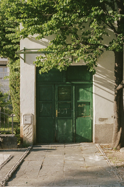 A wide, lush green door on a beige building under the setting sunlight.