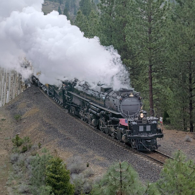 A color photo of a very large steam engine moving through a forest toward the lower right of the frame. An American flag flutters on the engine's right side. Very large billows of steam boil up and toward the upper left of the frame.