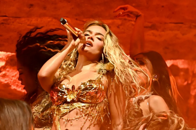 Performance shot of Karol G at Coachella 2026, wearing gold-plated bikini top featuring orchid centerpiece and branch/leaf motif. Surrounded by dancers in front of red lit background.