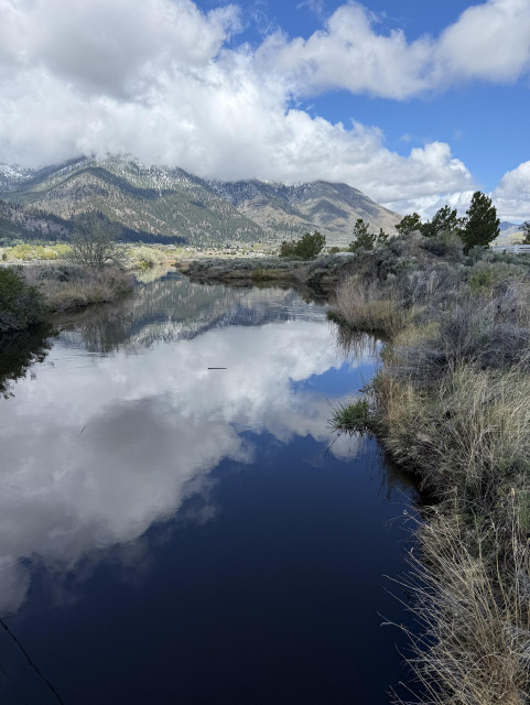 A river that reflects very blue sky and wild clouds leads to mountains where the clouds cast their shadows on. A narrow band of green pastures are at the bottom of the mountains.