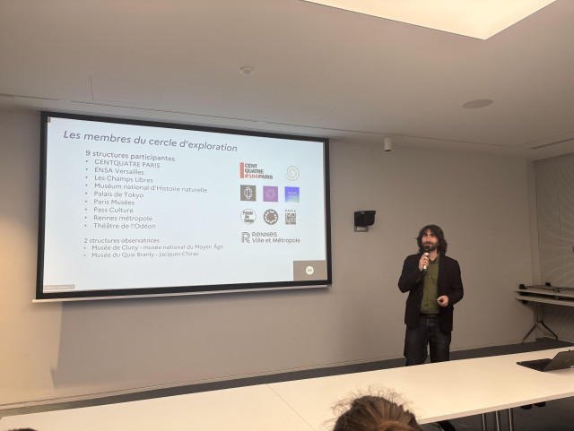 Photographie de Maxime présentant devant un écran qui affiche une diapositive intitulée « Les membres du cercle d’exploration » listant les 9 structures qui ont participé