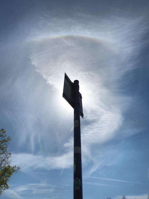 Foto des Himmels mit Cirremwolken. Die Sonne ist durch ein Schild verdeckt. Oberhalb der Sonne steht ein farbiger Ring, zentriert auf die Sonne.