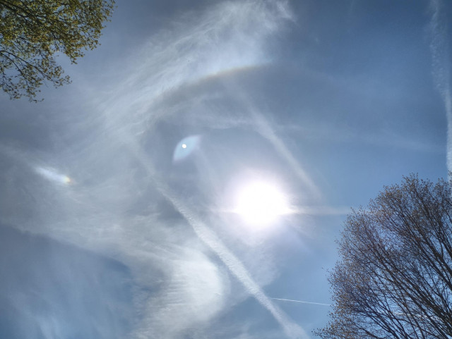 Foto des Himmels mit Cirruswolken. Oben links von der Sonne ein farbiger Ringteil, links von ihr ein heller, farbiger Fleck mit farblosem Schweif.