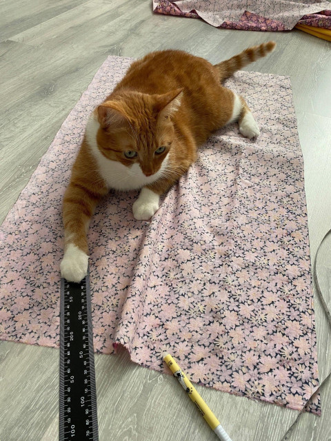 Orange and white cat lounging on a pink floral fabric spread across a wooden floor, with one paw resting on a black ruler beside a yellow fabric marker as if helping with sewing measurements.