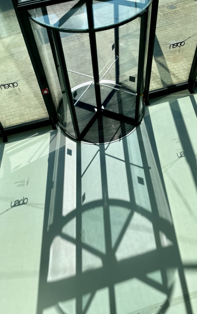 Farbiges Hochformat. Eine Drehtür von schräg oben fotografiert. Licht fällt durch die bodentiefen Fenster und die Drehtür auf den Boden. 