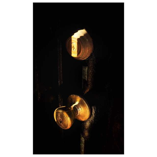 Closeup of a brass doorknob and the round brass keyhole cover above it, on a door with a coat of glossy black paint. A vertical strip of sunlight aligns perfectly with the keyhole and gives the brass components and surrounding paint a luminous glow, while leaving the rest of the image in shadow.