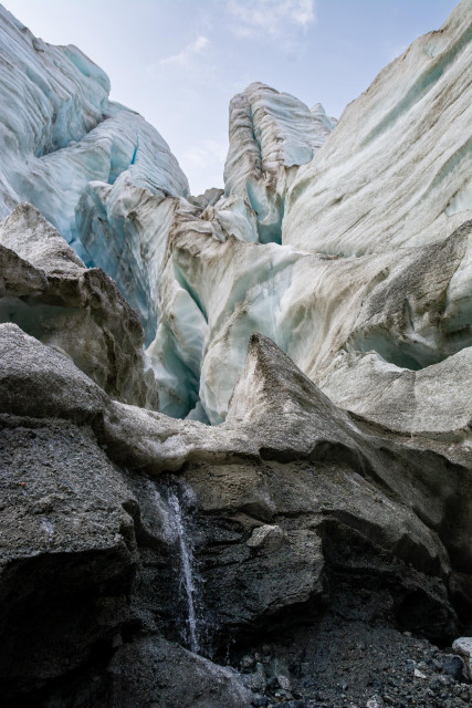 A low-angle upward view into the seracs of the Gabriel Icefall on the Gulkana Glacier, Eastern Alaska Range, Alaska. Towering walls of glacial ice rise on both sides of the frame, the surfaces streaked with sediment and smoothed by meltwater. Blue glacial ice is visible in the deeper portions of the serac walls at upper left. A thin trickle of meltwater falls from within the icefall down through dark sediment-covered ice and rock in the foreground. A pale blue sky with light clouds is visible at the top of the frame.