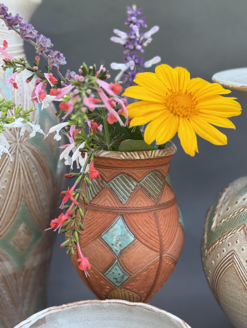 A decorative ceramic pot with a colorful geometric pattern contains a variety of fresh flowers, including a large yellow Mexican sunflower and smaller pink and purple salvia blooms. The pot is dark pink (maybe salmon) and light orange colors derived from natural clay. There are glossy aqua diamond-shaped accents, painted with glaze. The background is light gray and features more pottery in neutral tones.