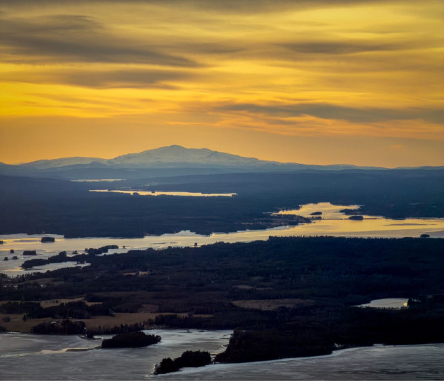A sweeping aerial panorama over the partly frozen Lake Storsjön at sunset. The sky is ablaze with deep gold and amber tones, reflecting in patches across the open water below. A snow-capped mountain — likely Oviksfjällen — rises in silhouette on the horizon. Dark forest and scattered islands fill the middle ground, with fields and smaller lakes visible in the foreground. The image conveys the vast, majestic scale of the Jämtland landscape.