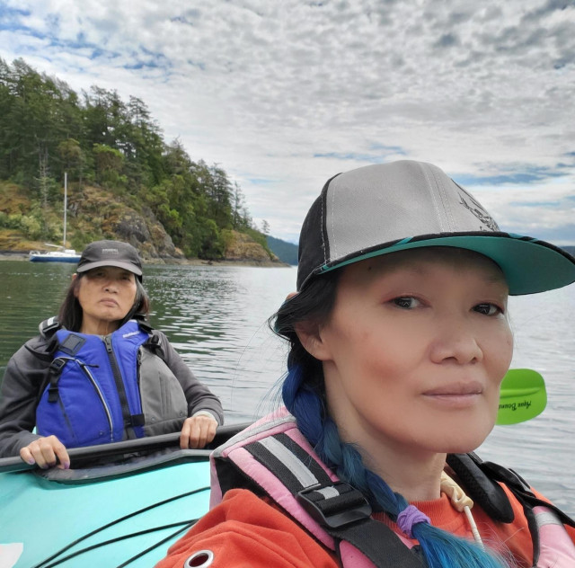 Me & Mom in a double kayak on the water with a wooded island and sailboat in the background under a cloudy sky.