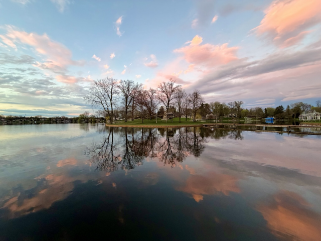 Photograph of a small island with bare trees on a still reflective lake at sunset with pink clouds above it also reflected in the water.