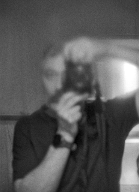 A blurred self-portrait seen through a fogged mirror. The figure holds a camera up to their face, but their features dissolve into soft shapes and grain. Moisture on the glass diffuses the light, turning the scene into a hazy, almost dreamlike silhouette. The body is partially visible—an arm raised, a dark shirt—but identity slips away, reduced to gesture and presence rather than detail.