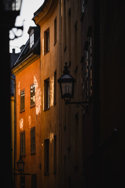 A vertical photograph showing the curving facade of a row of three-story buildings in an alley in Stockholm old town. The buildings are plastered/painted in an ochre colour and are indirectly illuminated by sunlight reflecting off of the windows of the building opposite. The lighting is brightest far back in the alley, with the foreground mostly in shadow. 

The building on the other side is just visible as a blurred sliver of a silhouette along the left edge of the frame.