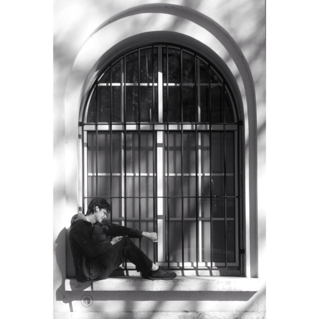 Black and white street photography taken in Paris showing a teenager sitting on the edge of a window. The window has the shape of D letter, turned 90° up. Some shades from a tree create a pattern on the image.