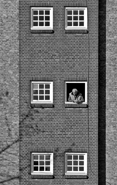 A photo of a tall brick building with three sets of white-framed windows. A person is leaning on the windowsill of the middle window, looking out. 