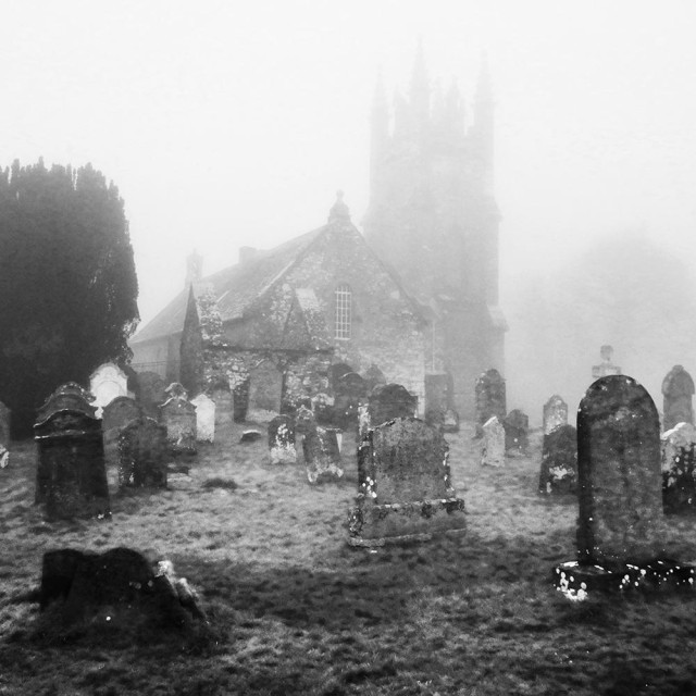 Atmospheric black and white photo, depicting gravestones in the foreground and the misty outline of a church with a bell tower behind.