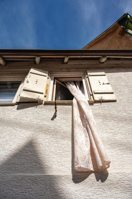 In dieser Nahaufnahme weht ein langer, heller Vorhang aus einem weit geöffneten Fenster eines älteren Hauses mit weißer Putzfassade. Der Stoff des Vorhangs ist cremefarben und mit einem zarten, gold- oder ockerfarbenen Muster aus großen Rosenblüten bedruckt. Er hängt fast über die gesamte untere Bildhälfte hinab und wirft einen deutlichen Schatten auf die raue, strukturierte Außenwand. Das Fenster selbst wird von zwei hellen Klappläden aus Holz flankiert, die ebenfalls leicht geöffnet sind und Abnutzungserscheinungen wie kleine Risse und verwitterte Stellen zeigen. Über dem Fenster schließt das Dach mit einem dunklen Dachüberstand und sichtbaren Holzbalken ab, während im Hintergrund ein strahlend blauer, wolkenloser Himmel zu sehen ist. Die Perspektive von unten nach oben.
