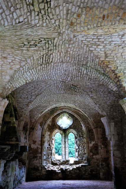 An ancient room within a ruined abbey with a complex vaulted ceiling composed of small pieces of stone. At the end of the room is a church-like window with a quatrefoil at the top and two tall arched lights below. The stonework around the window is intact at the side and top but broken at the bottom, though obviously consolidated. Out of the window can be seen a grassy area with a large tree.