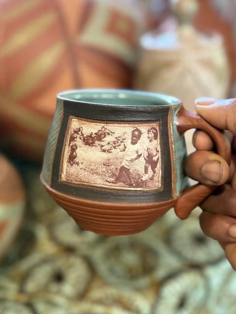 My hand holding a  ceramic mug featuring a vintage photo of two children surrounded by ducks. The mug has a textured pattern and a distinctive design, with a glossy aqua interior and an earthy tone on the exterior. Background features other decorative pottery.