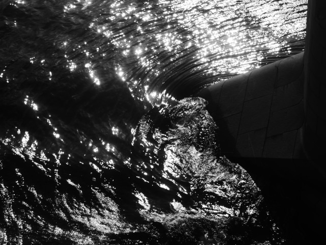 Черно-белая фотография: вид с моста через Неву вниз на воду, которая кружится и пузырится у одной из каменных опор. На воде играют блики от солнца.