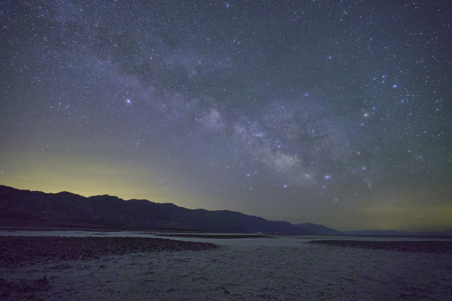 A wide and dense band of stars across the night sky over a cracked desert landscape. The sky on the horizon is lit from distant city lights.