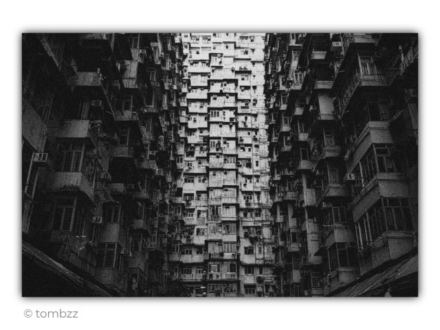A black and white, heavily grained photograph showing the famous Yick Cheong Building (known as the Monster Building) in Hong Kong. The low-angle shot reveals a monumental, raw wall of windows, balconies, and air conditioning units that create a dense, almost claustrophobic structure. The side wings of the buildings are shrouded in shadow, contrasting with the brighter facade in the center, giving the whole scene a dark, brutalist character. The distinct grain texture emphasizes the rawness and industrial atmosphere of this urban jungle.