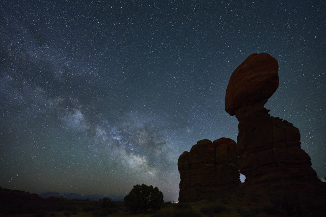 A wide and dense band of stars across the night sky. In the foreground is a stone tower that looks like a large rock is balanced at the top.