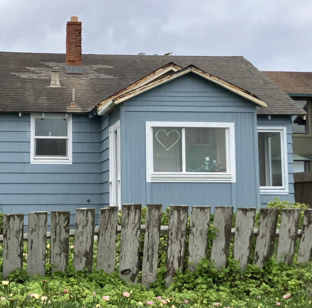 A basic window in a house painted a blue-gray. Near the top of one side of the window, in white, is a simple heart. 