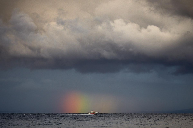 A small white-and-red boat moves across a dark, turbulent sea. In the distance, a section of rainbow shines beneath a grey, misty rain curtain fading into the horizon. A mass of brooding, stormy clouds looms over the entire scene, hanging heavily in the sky. The low horizon stretches towards a faint, dark coastline, barely visible in the distance. Rain streaks down from the clouds, blurring the boundary where the dark water meets the shadowed sky.